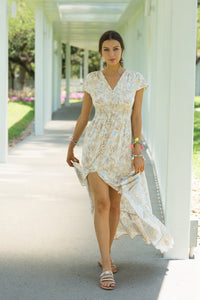 Model wearing white maxi dress with boho floral print in beige and blue with v neck and buttons all the way down to a front slit and cap sleeves. 