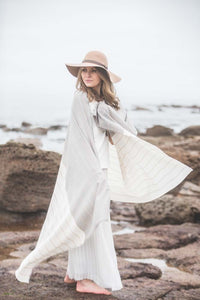 Model by seaside wearing cream and grey striped lightweight blanket scarf and floppy hat